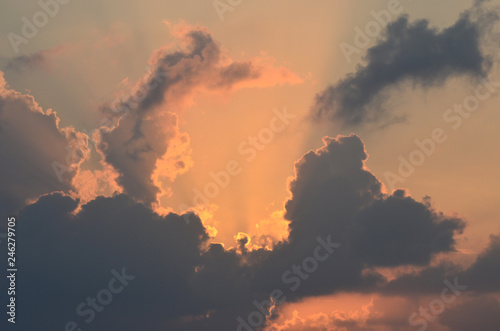 The rising sun is hidden by morning clouds. The clouds range from dark grey to light orange in colour. Streams of sunlight spread out from behind the clouds into a pale sky.