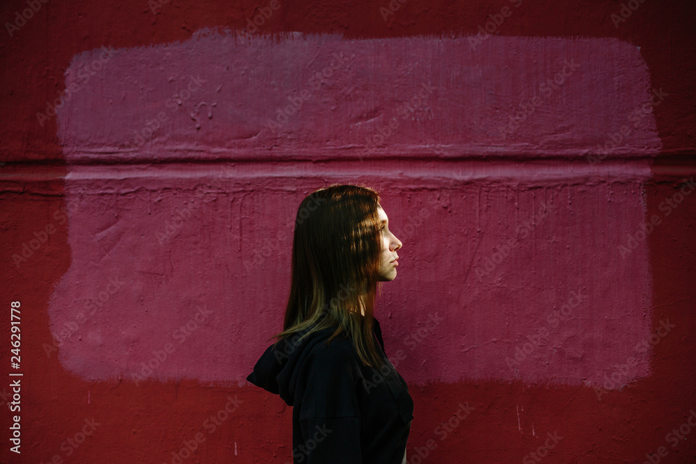 Outdoor city portrait of Young teen girl wearing black hoody, red wall on background, sunlight on the face