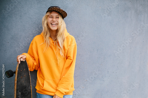 City portrait of positive young female wearing orange hoody and baseball cap holding skateboard. Grey blue wall on background