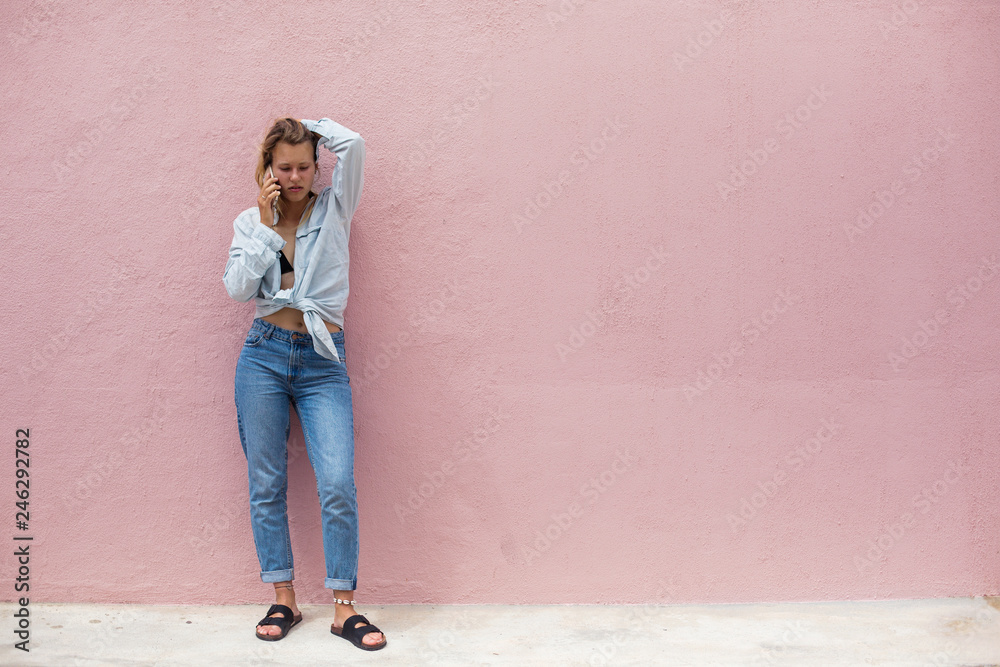 Young sad girl talking by mobile phone, standing outdoor with pink wall on background. Space for text