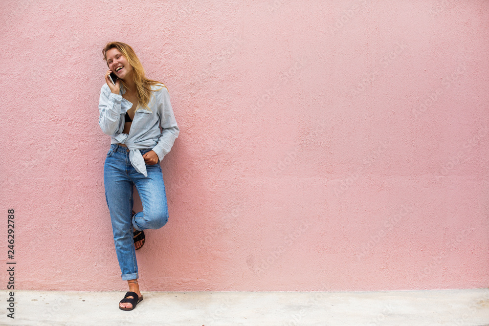 Young happy girl talking by mobile phone, standing outdoor with pink wall on background. Space for text