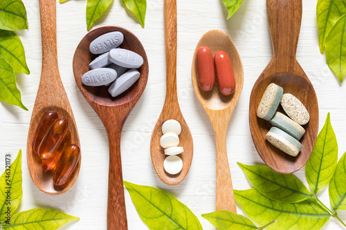 Variety of vitamin pills in wooden spoon on white background with green leaf, supplemental and healthcare product, flat lay surface photo