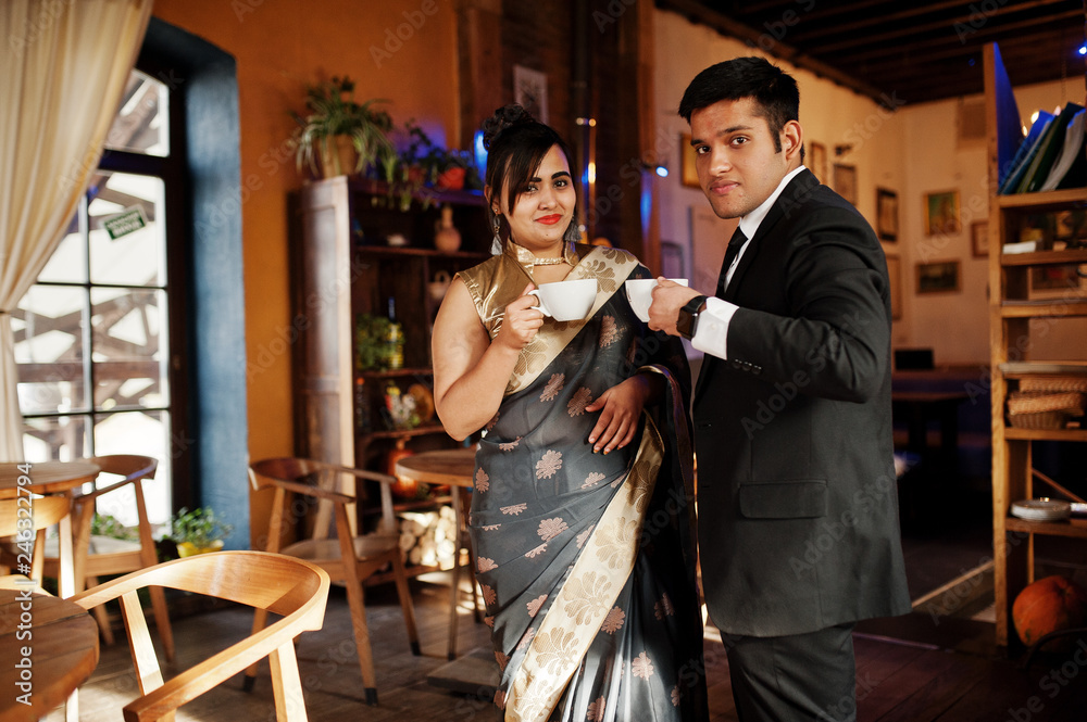 Elegant and fashionable indian friends couple of woman in saree and man in suit sitting on cafe and drinking tea.