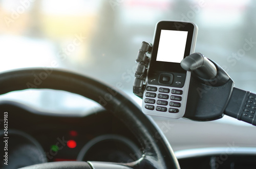 Mobile phone with blank screen in a robotic hand on a car steering wheel and dashboard background. Handsfree driving or automatic driver assistant concept.