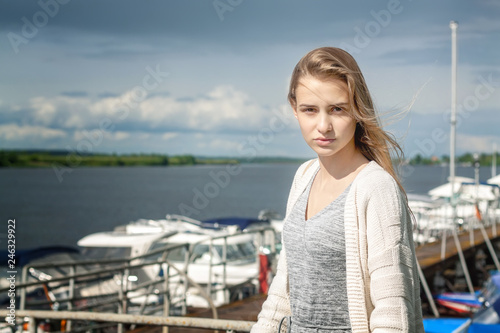 A beautiful girl stands alone on the pier against the background of the watery surface and thunderclouds extending beyond the horizon. The wind spreads her blond hair. © Dmitry