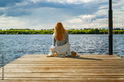 A beautiful girl is sitting alone on the river pier against the backdrop of thunder clouds and looks into the distance. View from the back. The wind develops her curls of red hair. 