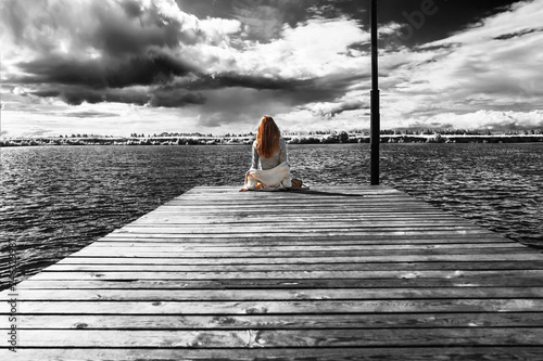 A beautiful girl is sitting alone on the river pier against the backdrop of thunder clouds and looks into the distance. View from the back. The wind develops her curls of red hair. 