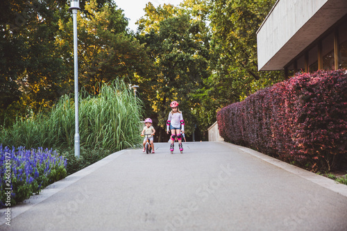 Two little girls rides on rollers and runbike in summer park. Children wearing protection pads and safety helmet for safe ride. Active outdoor sport for kids. Siblings have competition photo