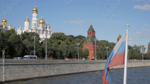 Russian flag on boat in front of red brick Kremlin walls, towers and white churches with golden domes on summer day in Moscow, Russia