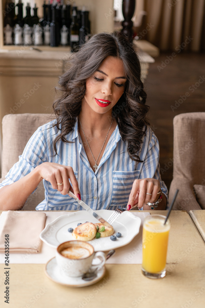 Beautiful young woman cuts pancakes with a knife, enjoying coffee cappuccino or latte in a glass cup with foam. Light breakfast near window in a cafe. Brunette with red bright lipstick Beautiful young woman cuts pancakes with a knife, enjoying coffee cappuccino or latte in a glass cup with foam. Light breakfast near window in a cafe. Brunette with red bright lipstick