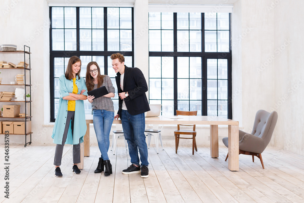 Fototapeta premium Creative teamwork. Positive office workers - two women and man stand near large wooden table and look at the tablet discussing new project on the background of large windows in office