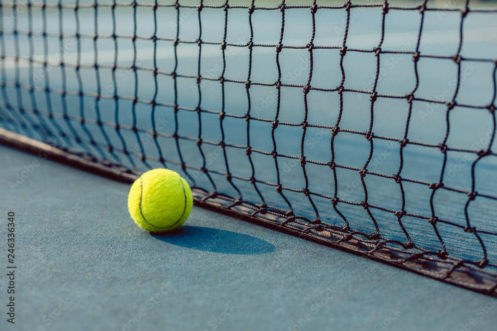 Close-up of a fluorescent yellow ball on blue acrylic surface in front of the net of a modern professional tennis court