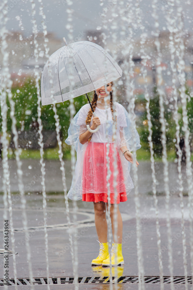 Naklejka premium Young pretty girl with two braids in yellow boots and with transparent umbrella stands near fountain.