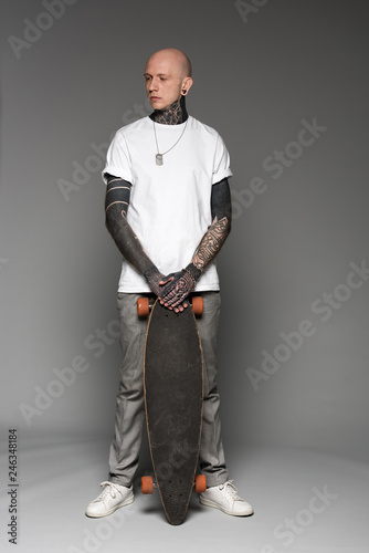Full length view of handsome bald tattooed man in white t-shirt standing with skateboard and looking away on grey
