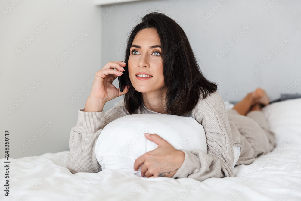 Photo of adorable woman 30s using mobile phone, while lying in bed with white linen in bright room Photo of adorable woman 30s using mobile phone, while lying in bed with white linen in bright room