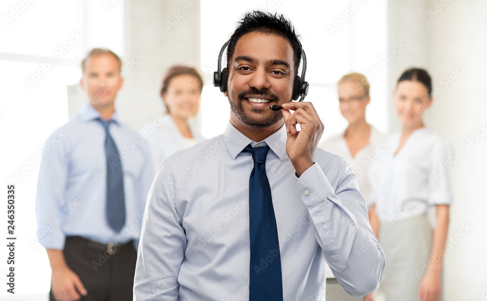 business, customer service and people concept - smiling indian businessman or helpline operator in headset over colleagues on background business, customer service and people concept - smiling indian businessman or helpline operator in headset over colleagues on background