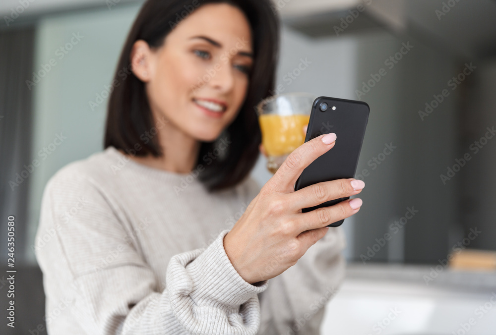 Image of satisfied woman 30s drinking orange juice and using mobile phone, while resting in bright modern room Image of satisfied woman 30s drinking orange juice and using mobile phone, while resting in bright modern room