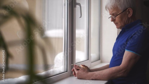 Confident senior man standing near the window while wearing glasses and looking phone.