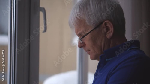 Closeup old man wearing glasses and surfing his phone near the window. Outside the window is snowy.