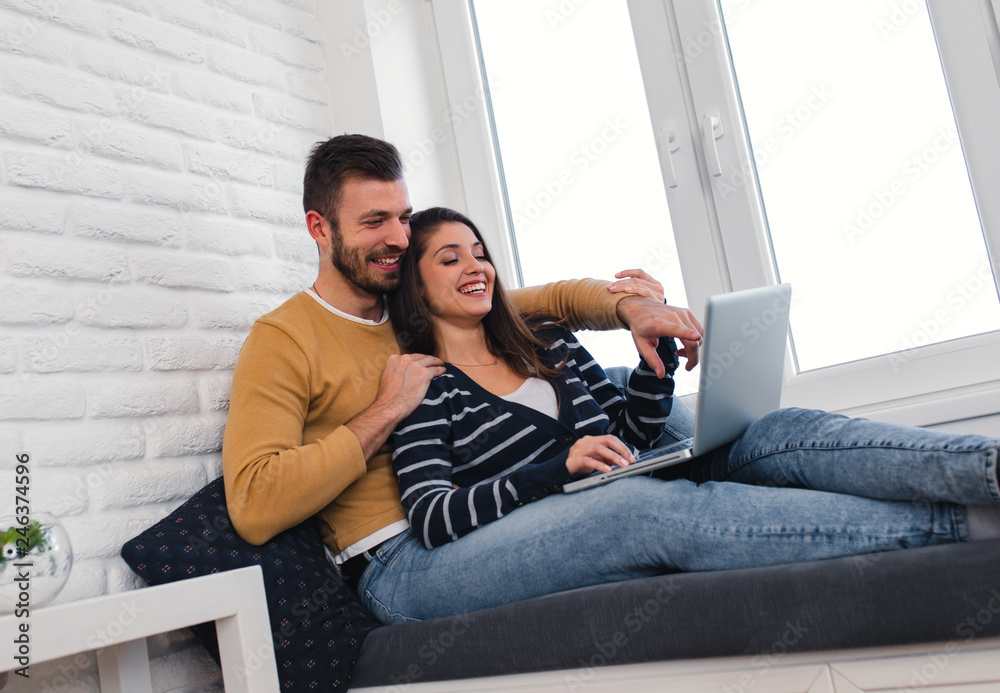 Obraz premium Young couple spending time together at home, they siting by the window and using at laptop.