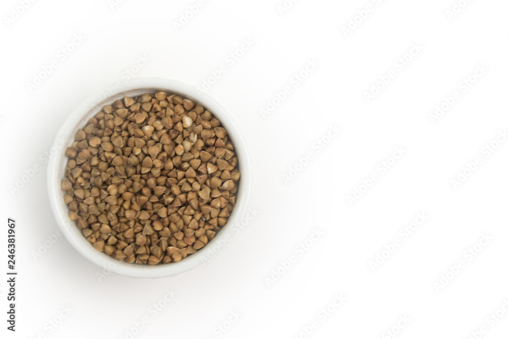 Dry buckwheat in white ceramic bowl isolated on grey table. Spilled buckwheat. Dry buckwheat in white ceramic bowl isolated on grey table. Spilled buckwheat.