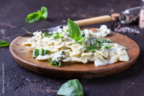 Pasta (farfalle) with creamy sauce with basil and gorgonzola cheese on a wooden plate. Dark brown concrete background. Copy space.