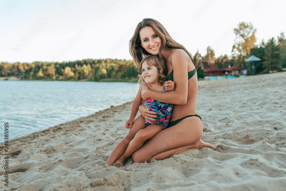 Obraz premium Beautiful and happy young woman mom hugging on shore of lake with sand, little child daughter, happy smiling, family resting in the summer resort on the shore. The concept of love and care parents.