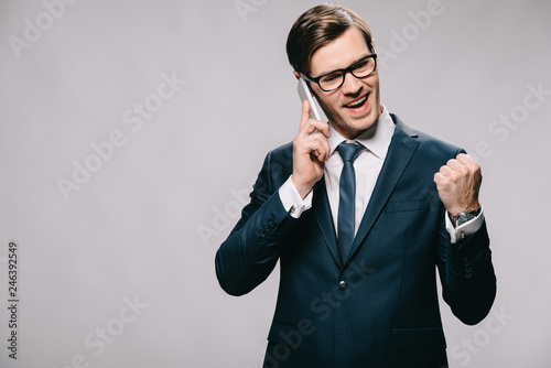 handsome businessman talking on smartphone and celebrating victory isolated on grey