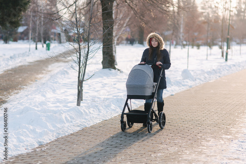 Beautiful mother walking in the park with her little baby in stoller. Woman dressed in blue jaket with hood and jeans. Warm boots photo