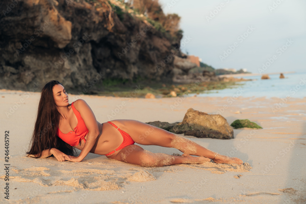 Sexy brunette girl in red swimsuit lies and sunbathes on beach with rocks.