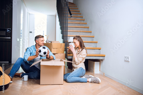 cheerful husband looking at wife drinking coffee while sitting on floor with football