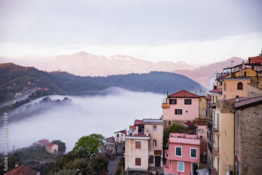 Panoramic top view of Alps mountains in fog and clouds, valley in sunrise. Medieval buildings of Perinaldo town, Liguria, Italy