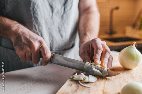 Fotografie, Obraz Rough male hands thinly chop a white onion with a knife on a wooden board - in a