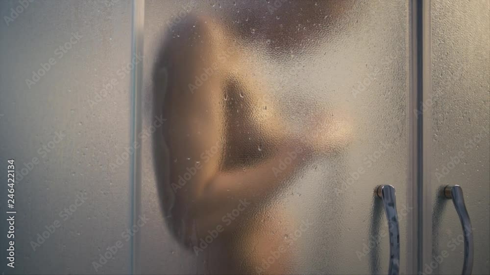 Unrecognizable silhouette of young fit woman in shower, focus on waterdops on glass