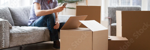 Close up carton boxes with belongings man sitting on sofa hold phone on background, wait call to delivery service moving at new house concept, banner for website header design with copy space for text
