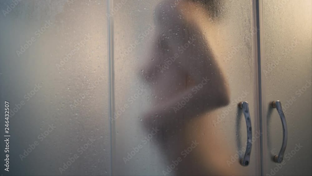 Unrecognizable silhouette of young fit woman in shower, focus on waterdops on glass