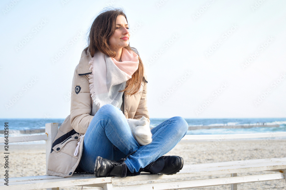 A young beautiful woman sits on a bench alone at sea and looks into the distance.