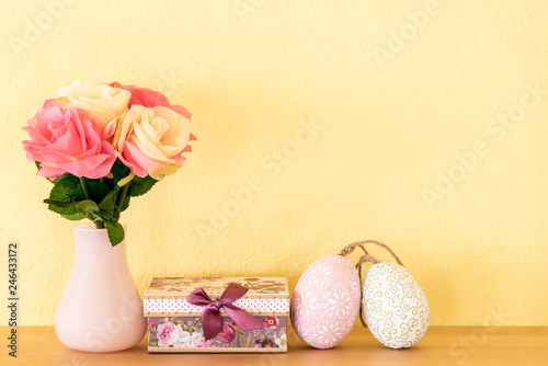 Rosen in der Vase mit Geschenk und zwei Ostereier vor einem gelben Hintergrund