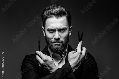 Portrait of bearded hipster male barber holds sharp scissors and a blade isolated on gray backgrund photo