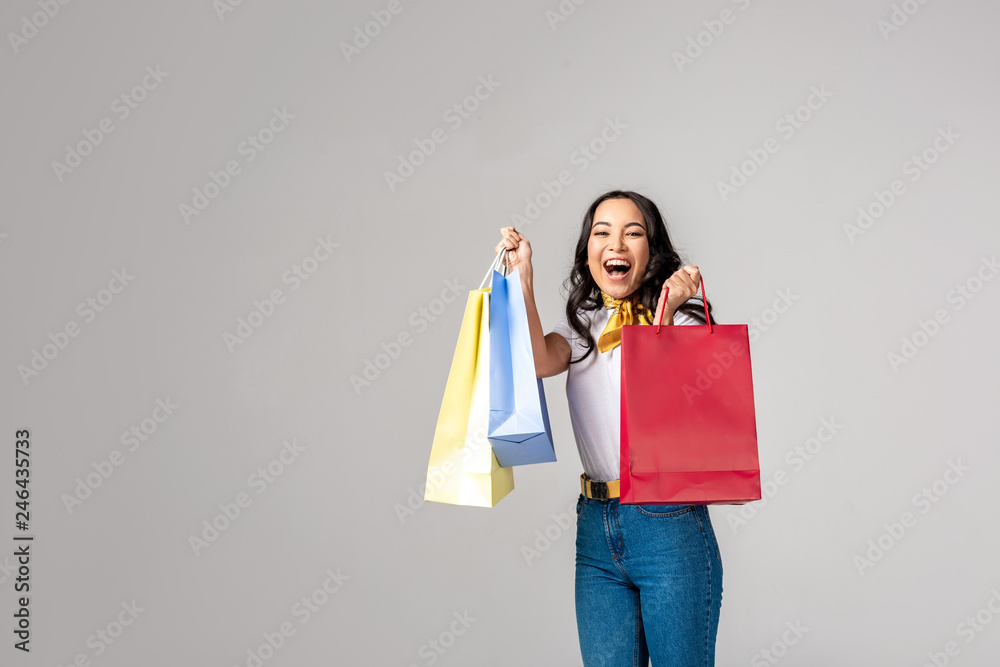 Smiling asian woman holding colorful shopping bags on raised hands and looking at camera isolated on grey