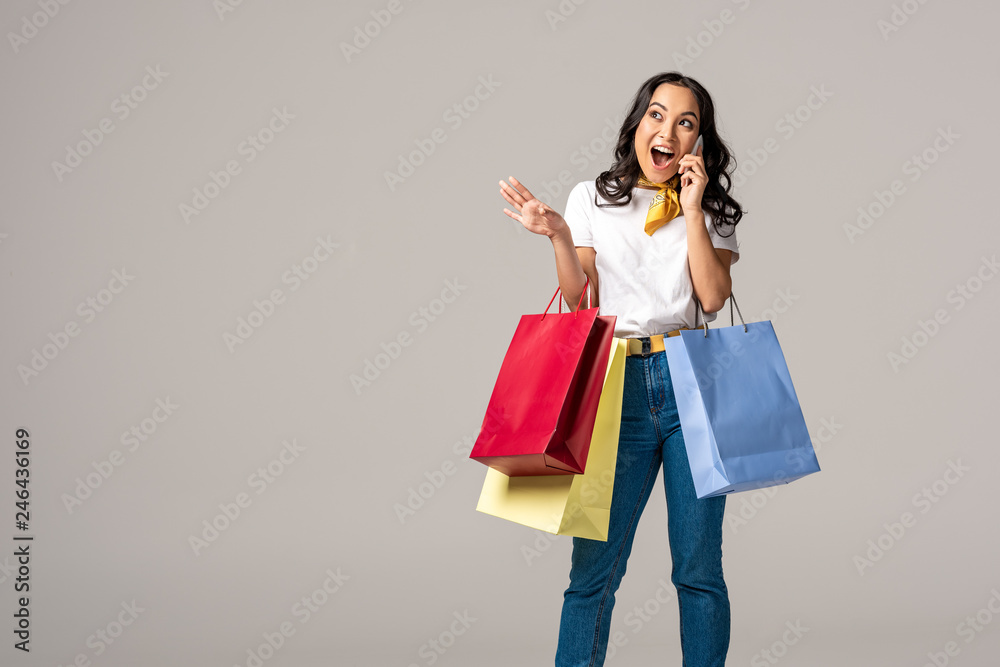 Excited young asian woman holding colorful shopping bags and emotionally talking on smartphone isolated on grey