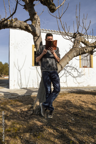 Young man standing and reading a book leaning on a tree in the outside of a rural house of the countryside. Scene of a beautiful and sunny day.