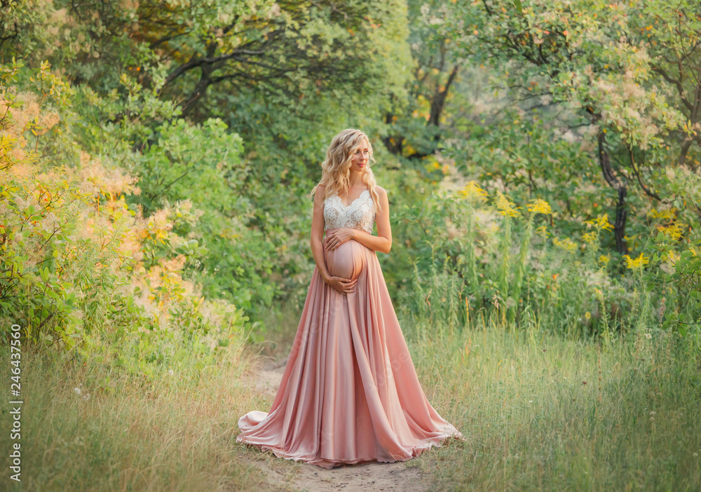 young sweet pregnant girl with light curly hair hugs and holds her tummy, standing in a fairy forest in cold shades. dresses a long elegant satin silk pink dress with a lace top. art processing photo