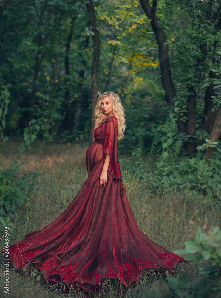 Fototapeta premium young pregnant woman with blond curly hair in long light flapping red scarlet dress, standing in the forest, holding, hugging her tummy. like a fairy magnificent queen. art autumn dark halloween photo