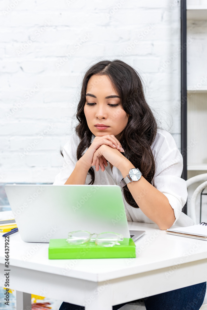 Tired asian freelancer looking at laptop screen and clutching hands under chin Tired asian freelancer looking at laptop screen and clutching hands under chin