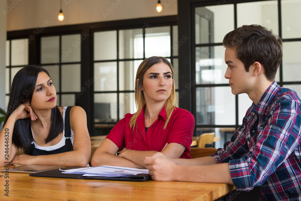Portrait of three young colleagues or students having discussion. Two women and man sitting at table and communicating. Planning or education concept Portrait of three young colleagues or students having discussion. Two women and man sitting at table and communicating. Planning or education concept