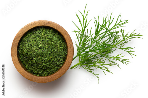 Slika na platnu Dried chopped dill in a dark wood bowl next to fresh dill leaves isolated on white from above