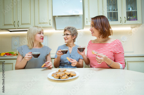 three grown women drink coffee with cookies in the kitchen