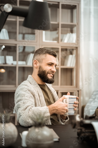 Smiling bearded man drinking hot tea while sitting at the desk