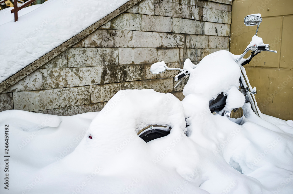 Fototapeta premium Winter in Russia. Motorcycle parked near the wall of the house is almost completely covered with snow and waiting for spring, and the beginning of the season for long journeys.